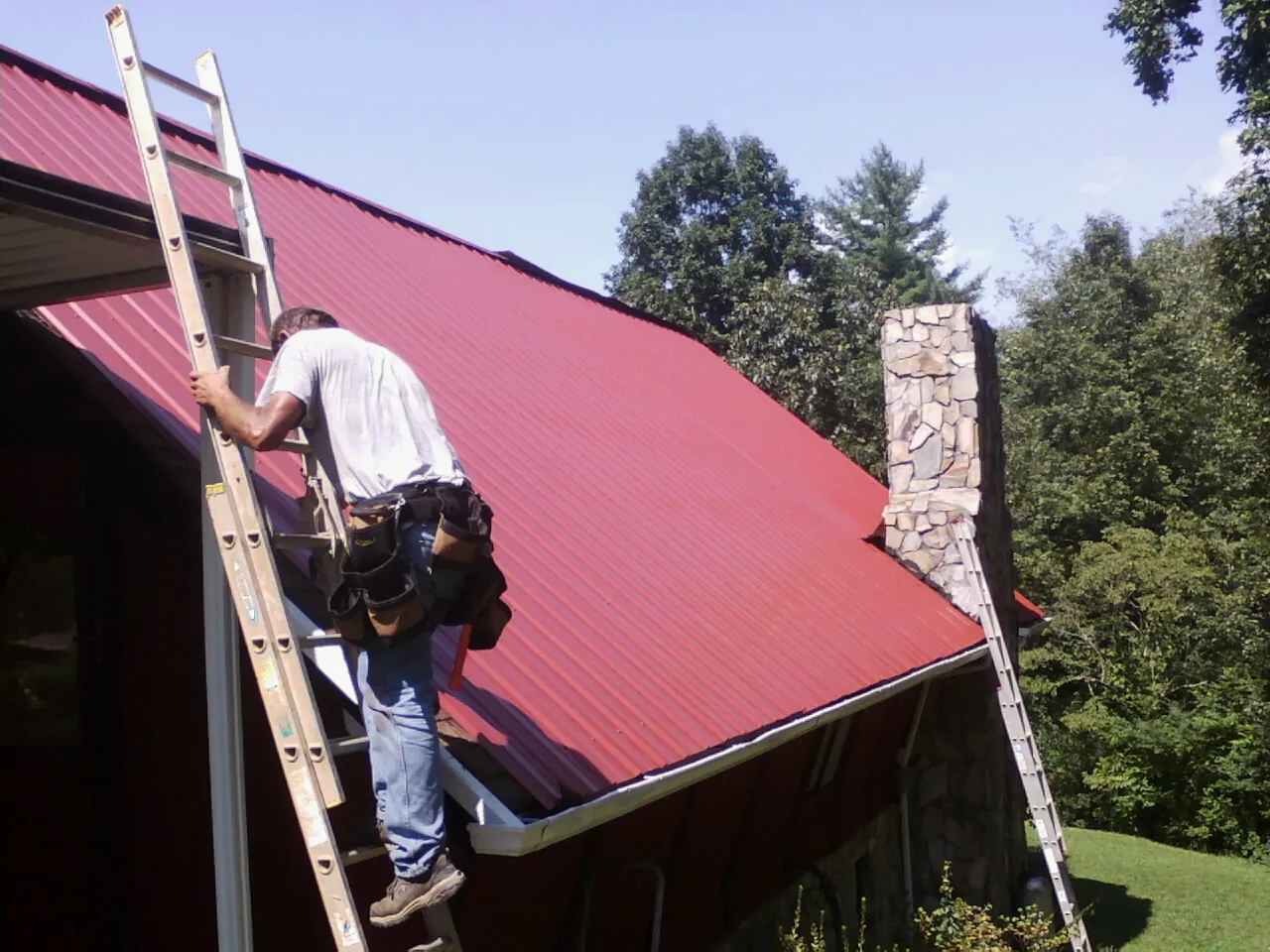 Roofer on a ladder beside a red metal roof and stone chimney — Soffit & Fascia Repair by Roofing Brothers in Block Island